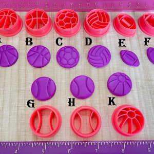 May include: A set of 11 pink and purple plastic cookie cutters shaped like different sports balls, including basketball, soccer ball, football, baseball, and volleyball. The cutters are arranged in two rows on a wooden surface.