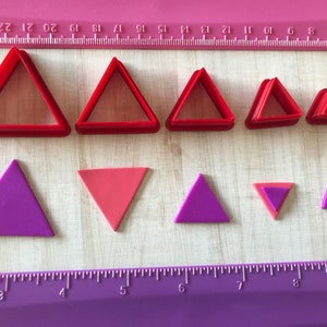 May include: A collection of red triangle-shaped cookie cutters in various sizes, alongside purple and pink triangles. A purple ruler with centimeter markings is at the bottom. The cutters are on a light wood-grain surface.