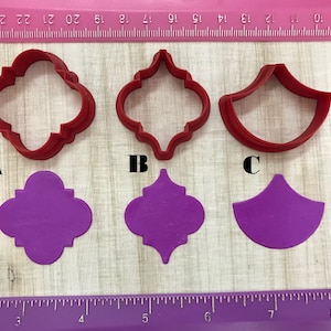 May include: Three red plastic cookie cutters in the shapes of a four-leaf clover, a quatrefoil, and a half-circle. Below the cutters are three purple fondant cutouts in the same shapes.