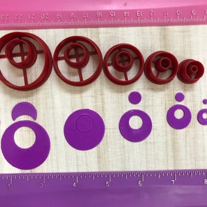 May include: A set of eight red and purple plastic cookie cutters in various sizes. The cutters are arranged in two rows on a wooden surface with a ruler.