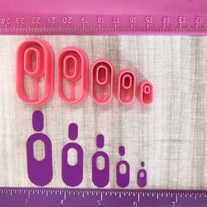 May include: A set of pink plastic oval-shaped cookie cutters in various sizes, arranged on a wooden surface. Below are matching purple cutouts. A pink ruler with centimeter markings is in the background.