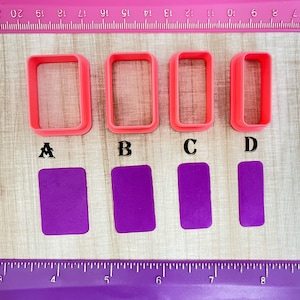 May include: Four pink plastic rectangle cookie cutters labeled A, B, C, and D. Below the cutters are four purple rectangles of fondant, each cut with the corresponding cutter.