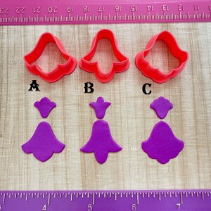 May include: Three red plastic cookie cutters shaped like bells, with the letters A, B, and C underneath. Three purple fondant bells, one for each cutter, are shown below.