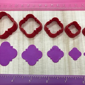 May include: Set of six red plastic cookie cutters in various sizes, shaped like a four-leaf clover with rounded edges. The cutters are shown next to five purple fondant cutouts of the same shape.