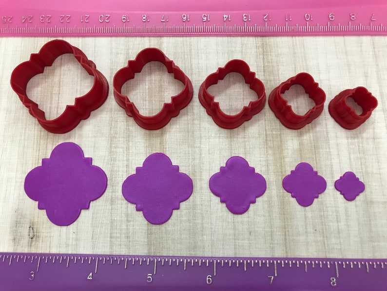 May include: Set of six red plastic cookie cutters in various sizes, shaped like a four-leaf clover with rounded edges. The cutters are shown next to five purple fondant cutouts of the same shape.