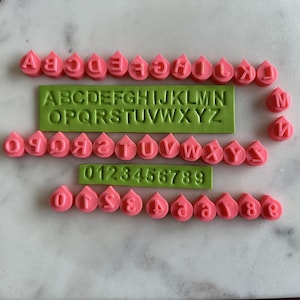 May include: A set of pink and green alphabet and number stamps. The stamps are shaped like raindrops and have the letters A through Z and the numbers 0 through 9.