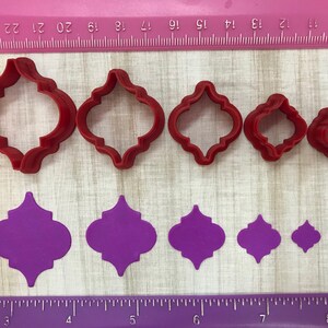 May include: Set of six red plastic cookie cutters in various sizes shaped like a quatrefoil, and six purple fondant cutouts in the same shape.