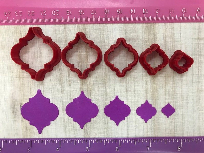 May include: Set of six red plastic cookie cutters in various sizes shaped like a quatrefoil, and six purple fondant cutouts in the same shape.