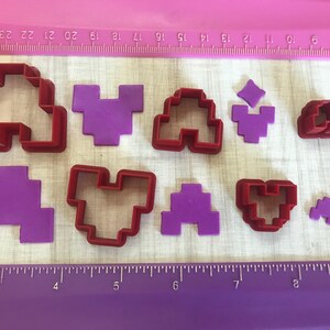 May include: A set of eight red and purple plastic cookie cutters in various shapes, including hearts, arches, and squares. The cutters are arranged on a white surface with a purple ruler.