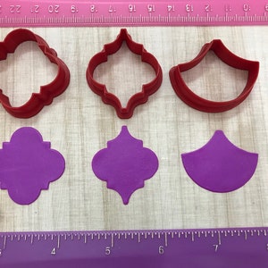 May include: Three red plastic cookie cutters and three purple fondant cutouts. The cutters are shaped like a quatrefoil, a quatrefoil with a center point, and a half-circle. The cutouts are the same shapes.