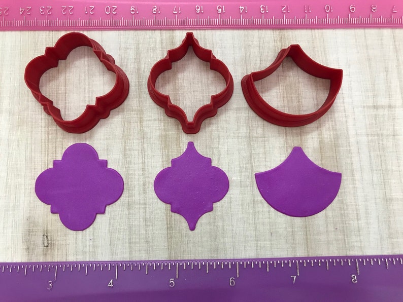 May include: Three red plastic cookie cutters and three purple fondant cutouts. The cutters are shaped like a quatrefoil, a quatrefoil with a center point, and a half-circle. The cutouts are the same shapes.