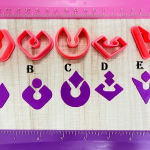 May include: A collection of five red plastic earring-shaped cookie cutters and five purple earring cutouts. The cutters are in various shapes, including a hexagon, heart, and a V-shape. A ruler is visible in the background.