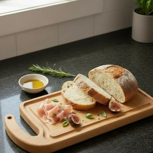 May include: A wooden cutting board with sliced bread, prosciutto, figs, and olive oil. The bread is partially sliced, with two slices visible. The prosciutto is arranged in a small pile, and the figs are cut in half. A sprig of rosemary is also present.