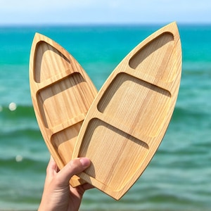 Puede incluir: Dos bandejas de servir de madera con forma de tablas de surf. Las bandejas son de madera marrón claro y tienen un acabado natural de madera. Las bandejas están apiladas una encima de la otra.