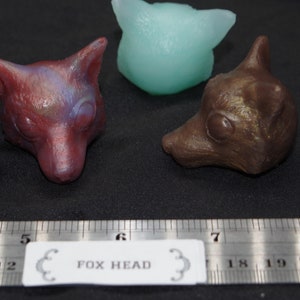 May include: Three fox head sculptures, one is a teal blue, one is a reddish brown with purple and blue accents, and the other is a bronze color. The sculptures are on a black surface with a ruler that says "FOX HEAD" in the middle.