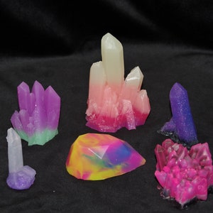 May include: A collection of six colorful crystal-shaped soap bars. The soaps are in various shades of pink, purple, green, and yellow. The soaps are arranged on a black surface.
