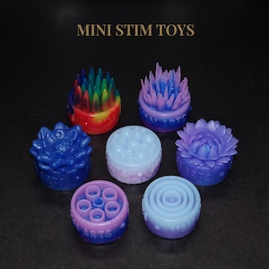 May include: A collection of seven miniature, colorful stim toys. The toys are various shapes, including flower and dome designs, and feature a range of colors like blue, purple, and red. The text "MINI STIM TOYS" is displayed at the top.
