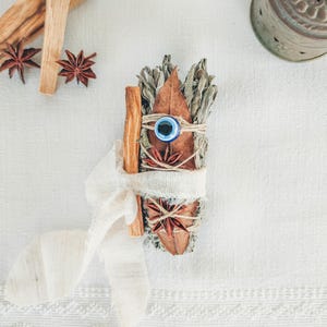 Może przedstawiać: Biały materiał z wiązką suszonych ziół, patyczkiem cynamonu i anyżem gwiazdkowym. Wiązka jest związana białą wstążką i niebieskim amuletem.