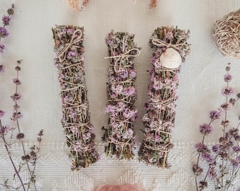 May include: Three bundles of dried herbs tied with twine, each bundle is wrapped with purple flowers. The bundles are arranged in a row on a white surface.