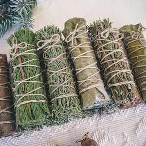 Op de afbeelding: Een verzameling van zes wierookstokjes, gebonden met touw. De wierookstokjes zijn gemaakt van gedroogde kruiden en bladeren, waaronder cederhout, salie en palo santo.