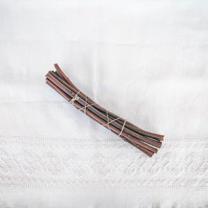 以下が含まれることがあります： 紐で結ばれた細い茶色の小枝の束。小枝は湾曲した形に配置され、白い布の表面の上に置かれています。