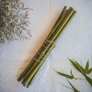 Op de afbeelding: Een bundel groene bamboestengels samengebonden met touw. De bamboe heeft een lichtgroene kleur en een natuurlijke textuur. De bundel ligt op een wit oppervlak met een bloemstuk en bamboebladeren.