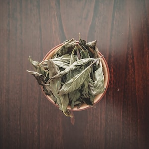 Puede incluir: Un cuenco marrón lleno de hojas verdes secas. El cuenco está sobre una superficie de madera marrón oscura.