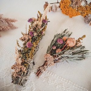 May include: Two bundles of dried herbs and flowers tied with twine. One bundle is a mix of yellow, green, and purple flowers, the other is a mix of green and brown herbs.
