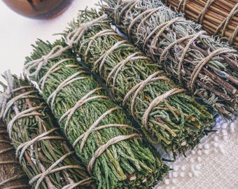 May include: A close-up of several bundles of dried herbs tied together with twine. The bundles are made of different types of herbs, including sage, rosemary, and lavender. The herbs are a natural green colour and are arranged in a row on a white surface.