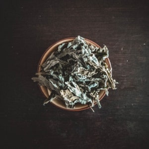 Op de afbeelding: Een close-up van een kom gevuld met gedroogde groene bladeren. De bladeren zijn in een stapel gerangschikt en hebben een lichtgroene kleur. De kom is bruin en heeft een glad oppervlak.