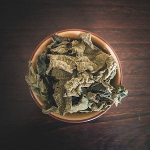 Op de afbeelding: Een bruine keramische kom gevuld met gedroogde groene bladeren. De bladeren zijn in een willekeurig patroon gerangschikt en lijken een soort kruid te zijn.