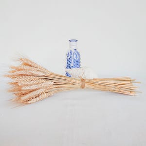 Puede incluir: Un manojo de espigas de trigo atadas con hilo, junto a una botella decorativa azul y blanca y un objeto blanco texturizado. El trigo es de color marrón claro, y la botella tiene una tapa blanca y un diseño azul.