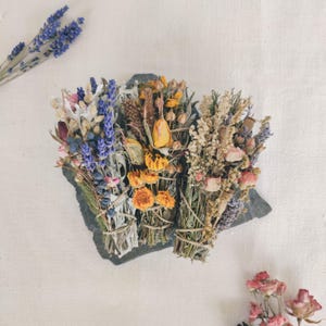 Può includere: Tre mazzi di fiori secchi, tra cui lavanda, fiori gialli e rosa, sono legati con spago. I mazzi poggiano su una pietra grigio scuro su uno sfondo bianco. Sono visibili anche un rametto di lavanda e alcune rose secche.