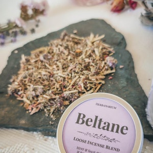 May include: A purple and silver tin of loose incense blend called "Beltane" with the text "HERBANARIUM" and "LOOSE INCENSE BLEND MINT & SAGE & YARROW & ROSES & ROSEMARY 10 GRAMS".