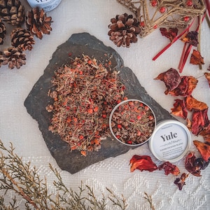 Puede incluir: Una lata plateada de mezcla de incienso Yule suelto con un diseño rojo y verde. La lata está abierta y el incienso se derrama sobre una superficie de pizarra gris oscuro. El incienso es una mezcla de hierbas secas y bayas.