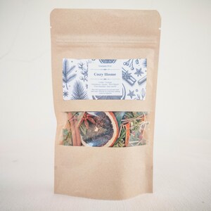 Puede incluir: Una bolsa de papel marrón con una ventana transparente que muestra una mezcla de rodajas de naranja secas, palitos de canela y otras especias. La bolsa tiene una etiqueta que dice "Cozy Home" y enumera los ingredientes: "Naranja, Canela, Anís estrellado, Pimienta roja, Agujas de pino, Hojas de laurel".