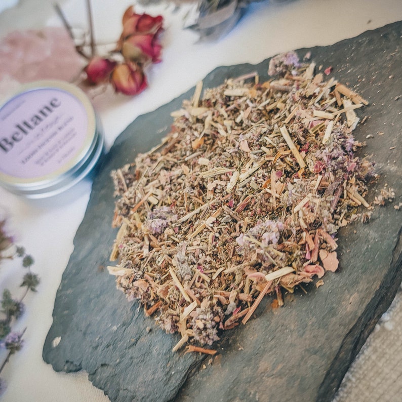 May include: A close-up of a pile of dried herbs and flowers on a dark gray slate surface. The herbs and flowers are a mix of brown, green, and purple.