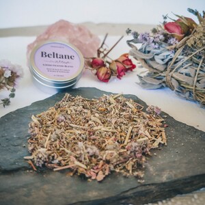 May include: A small tin of loose incense blend labeled "Beltane" with a pink and white label. The tin is sitting on a black slate with a pile of dried herbs and flowers.