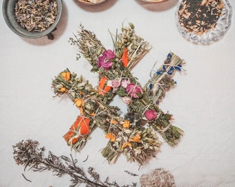 May include: Four bundles of dried herbs and flowers tied together with twine. The bundles are arranged in a star shape on a white surface. The bundles are decorated with dried flowers, including pink, purple, and white flowers.
