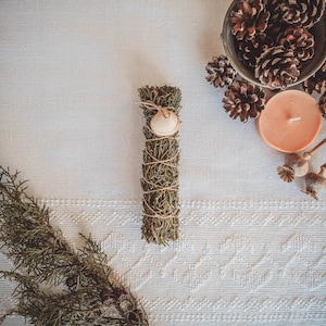 Puede incluir: Un manojo de salvia seca atado con cuerda y decorado con una concha marina. La salvia está sobre un fondo blanco con piñas y una vela.