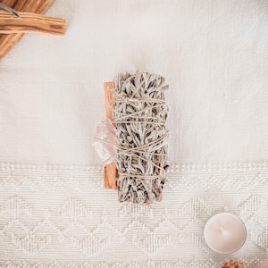 May include: A bundle of white sage smudge sticks tied with twine, wrapped around a wooden stick. A pink crystal is placed next to the bundle.