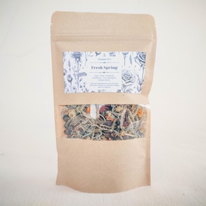 Peut inclure: Un sac en papier brun avec une fenêtre transparente montrant un mélange d'herbes et de fleurs séchées. L'étiquette sur le sac indique "Simmer Pot Fresh Spring" et liste les ingrédients : Sauge, Rose, Lavande, Romarin, Souci des champs, Baies de genévrier.