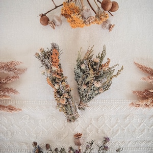 May include: Two bundles of dried flowers and herbs, one with a mix of green and brown, the other with orange and brown, tied with twine. The bundles are arranged on a white textured surface.