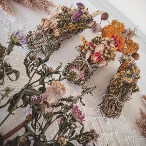 May include: Four bundles of dried herbs and flowers tied together with twine. The bundles are arranged on a white lace tablecloth. The bundles are made with a variety of dried flowers and herbs, including lavender, rosemary, and chamomile.