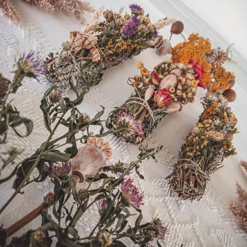 May include: Four bundles of dried herbs and flowers tied together with twine. The bundles are arranged on a white lace tablecloth. The bundles are made with a variety of dried flowers and herbs, including lavender, rosemary, and chamomile.