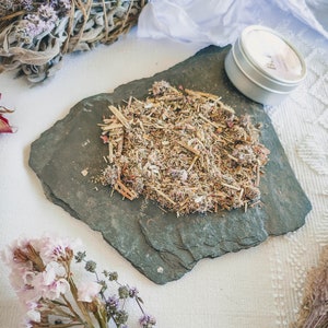 May include: A close-up of a small pile of dried herbs and flowers on a black slate plate. The herbs and flowers are a mix of brown, green, and pink. There is a small tin container with a white label that says "Bath" in the background.