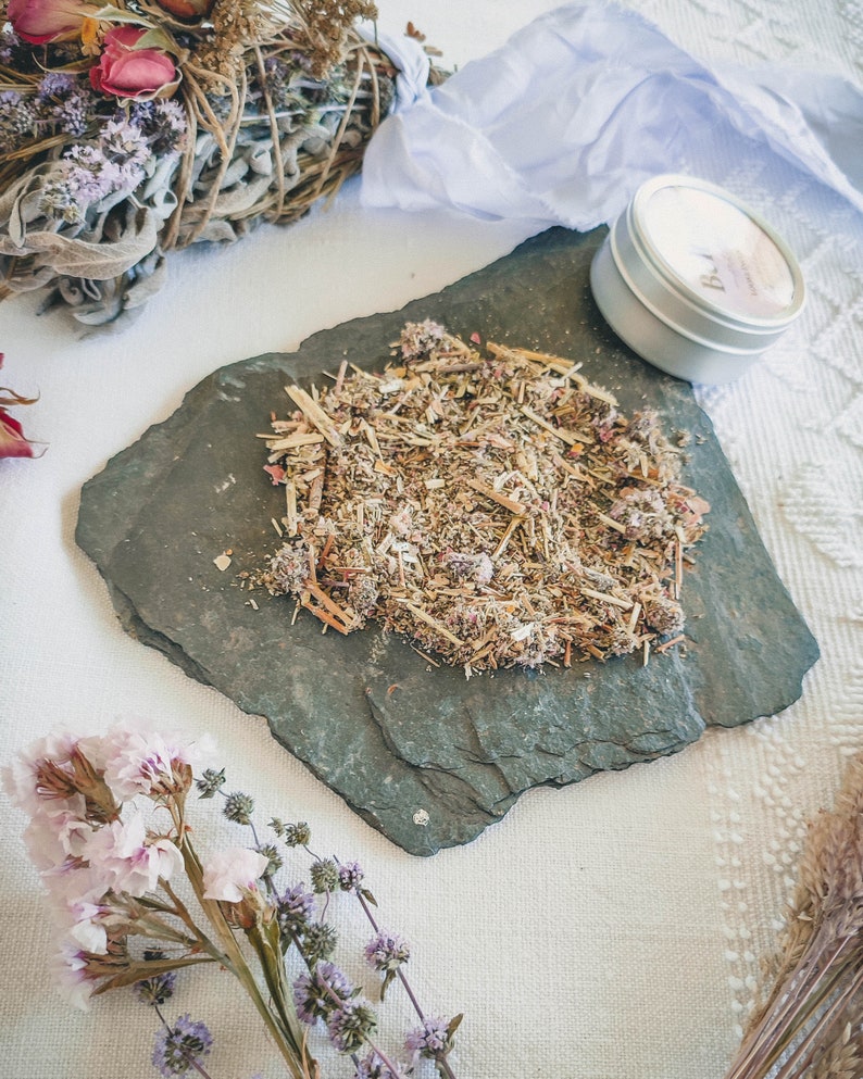 May include: A close-up of a small pile of dried herbs and flowers on a black slate plate. The herbs and flowers are a mix of brown, green, and pink. There is a small tin container with a white label that says "Bath" in the background.