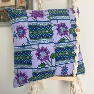 May include: A square throw pillow featuring a lavender fabric with a repeating pattern of purple flowers, green leaves, and geometric bands. The pillow is suspended by cream-colored macrame cord from a light wood dowel with fringe tassels.