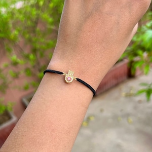 May include: A gold-coloured Hamsa hand charm bracelet with a pink centre stone and clear surrounding stones. The charm is attached to a black cord bracelet. The bracelet is on a person's wrist.