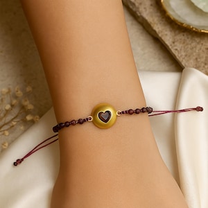 Puede incluir: Una pulsera dorada con una piedra preciosa en forma de corazón en el centro. La pulsera presenta una serie de pequeñas cuentas de color rojo oscuro y un cordón a juego. La piedra preciosa en forma de corazón es de color rojo intenso. La pulsera está en una muñeca.